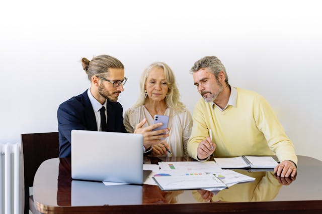 three people sitting in front of a laptop going over documents