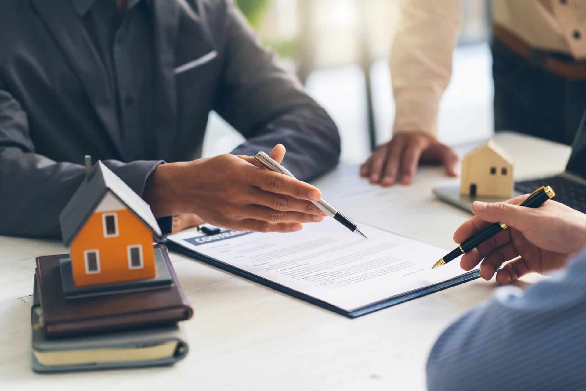 Hands signing a contract at a desk with a small house model.